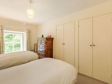 A bedroom with two beds a wooden chest of drawers a chair curtains on a window and closed closets at Gardeners Cottage in Bellingham