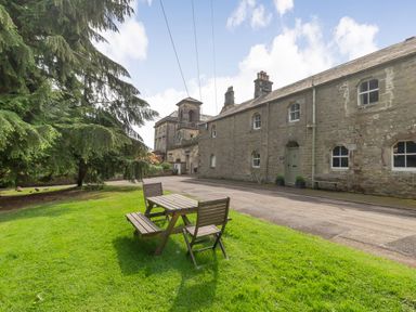 A wooden picnic table and bench on a grassy area near a stone building with windows and a clock tower at Gardeners Cottage in Bellingham