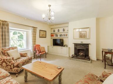 A living room with patterned armchairs a wooden coffee table fireplace and a built in bookshelf with a tv at Gardeners Cottage in Bellingham