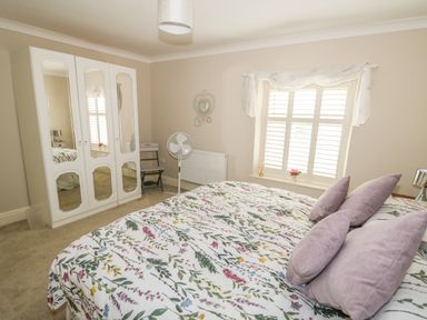 A bedroom with a bed with floral bedding and purple pillows a white wardrobe with mirrors a standing fan and a window with shutters at Bertelle in Grimston