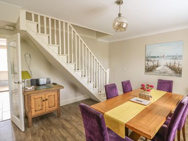 A dining room with a wooden table purple chairs a yellow table runner a painting on the wall a wooden cabinet under stairs and a hanging light at Bertelle in Grimston