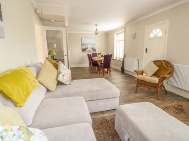 A living room with a grey sofa, a wicker chair with cushions, and a dining table with purple chairs at Bertelle in Grimston