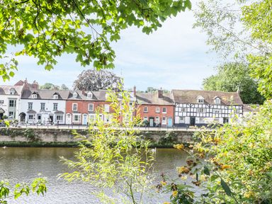 A river with old buildings and a riverside cafe across the water framed by tree branches and foliage at Town Mill in Bewdley
