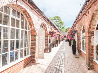 An outdoor corridor with brick walls large arched windows flowers in pots along the pathway and people walking at Town Mill in Bewdley