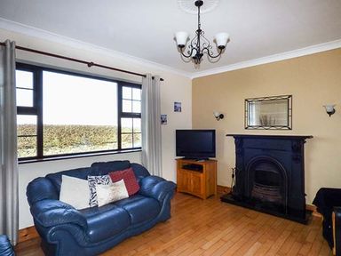 A living room with a blue couch a wooden floor a black fireplace a TV on a wooden stand and large window at Flynns Cottage in Quilty County Clare