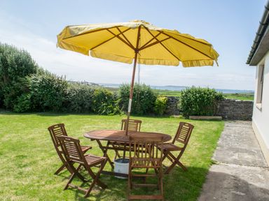 A wooden outdoor table with six chairs and a yellow umbrella on grass near bushes and a stone wall at Flynns Cottage in Quilty County Clare