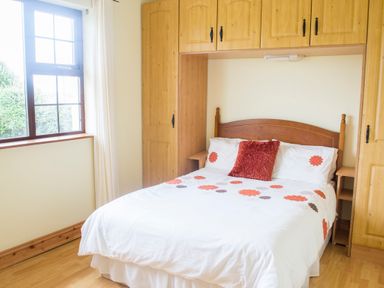 A bedroom with a double bed under wooden built-in cupboards and a window with white curtains at Flynns Cottage in Quilty County Clare