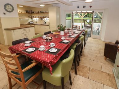 A dining area with a long table covered with a red polka dot tablecloth and chairs in a kitchen with cabinets and open shelves at Ty Llwyd in Nevern near Cardigan