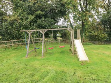 A wooden swingset with three swings and a slide in a grassy yard with trees and a wooden fence at Ty Llwyd in Nevern near Cardigan