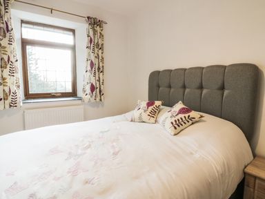 A bedroom with a bed with a gray upholstered headboard two patterned pillows a window with floral curtains and a wooden nightstand at Lower West Curry Farmhouse in Launceston
