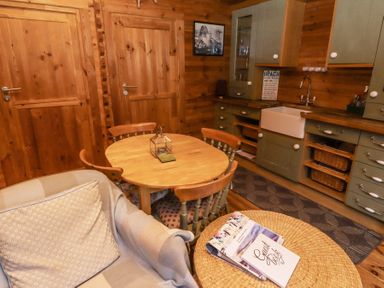 A kitchen with a wooden round table surrounded by four chairs a gray armchair with a cushion and throw a wicker table with books and a guest book on it green cabinets with a farmhouse sink and wood panel walls at Aurora Skies in Cresswell near Morpeth