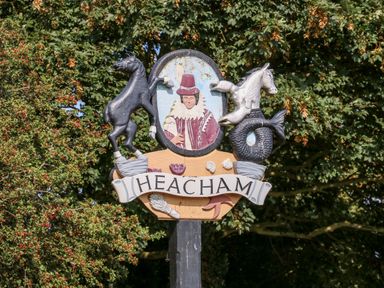 A signpost with the word Heacham featuring a man in historic clothing and two horse sculptures surrounded by trees