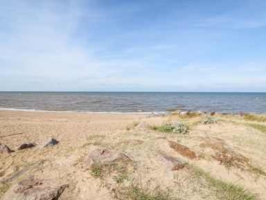 A sandy beach with rocks and grass patches by the sea under a blue sky at Carrstone Cottage in Heacham