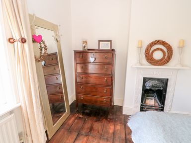 A bedroom corner with a wooden chest of drawers a full length mirror beige curtains and a white fireplace with decorative items at Carrstone Cottage in Heacham