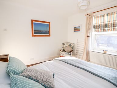 A bedroom with a bed pillows an armchair framed pictures and a window with striped curtain at Carrstone Cottage in Heacham