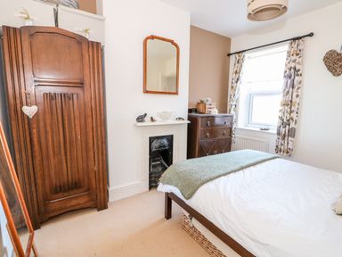 A bedroom with a wooden wardrobe a bed a small fireplace and a window with patterned curtains at Carrstone Cottage in Heacham