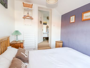 A bedroom with a wooden bed white bedding brown pillows a bedside table with a green lamp and a purple accent wall at Carrstone Cottage in Heacham