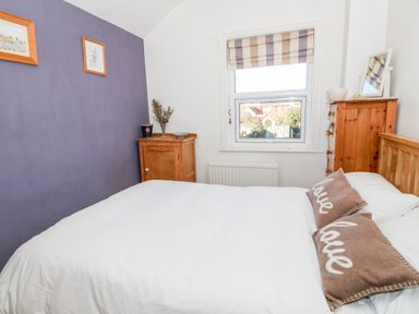 A bedroom with a bed white bedding brown pillows wooden furniture and a window with striped blinds at Carrstone Cottage in Heacham