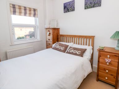A bedroom with a wooden double bed with two brown pillows on white bedding a wooden chest of drawers with a white bowl and a mirror and another wooden chest with a green lamp and a small bowl and two paintings above the bed at Carrstone Cottage in Heacham