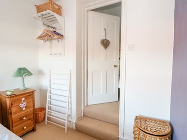 A bedroom corner with a wooden chest of drawers a green lamp a white door with a heart decoration hangers on a wall rack and a wicker basket at Carrstone Cottage in Heacham
