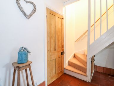 A hallway with a wooden door a small wooden stool with a blue fabric item on it a staircase with carpeted steps and white railings at Carrstone Cottage in Heacham
