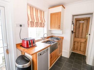 A kitchen with a red toaster on a wooden counter a stainless steel sink wooden cabinets a dishwasher a trash bin and a wooden door labeled bathroom at Carrstone Cottage in Heacham