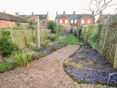 A garden with a gravel path bordered by plants and trees with brick houses in the background at Carrstone Cottage in Heacham