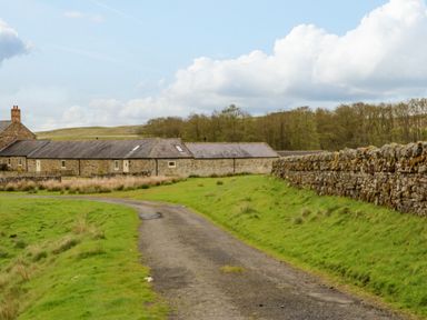 A country road leading to stone buildings with stone walls and grass fields at Gallow Law Cottage in Bellingham