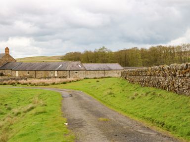 A rural stone building with a slate roof along a curved gravel road and a stone wall with grass and trees in the background at Shepherds Burn Cottage in Bellingham