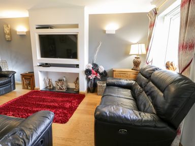 A living room with black leather sofas a red rug a wall mounted television and a wooden cabinet with a lamp at 6 Stad Yr Ysgol in Manod near Blaenau Ffestiniog