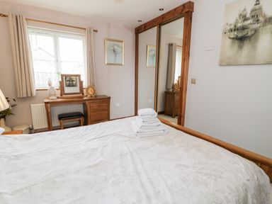 A bedroom with a bed covered with a white patterned duvet and folded towels on top a wooden dressing table under a window with beige curtains and a framed artwork on the wall next to a wooden wardrobe with mirrored sliding doors at 6 Stad Yr Ysgol Manod near Blaenau Ffestiniog