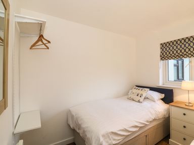 A bedroom with a single bed a nightstand with a lamp a window and a wall-mounted shelf with hangers at Keepers Cottage in Sidmouth