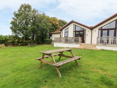 A grassy lawn with two wooden picnic tables in front of a building with large windows and a tiled roof at Keepers Cottage in Sidmouth