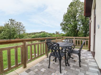 A patio with a black metal table and four chairs next to a wooden railing and green trees at Keepers Cottage in Sidmouth