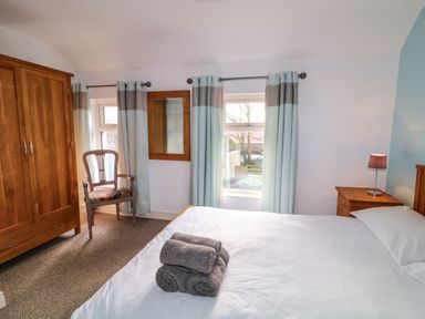 A bedroom with a bed with towels on it a wooden wardrobe a chair windows with curtains and a nightstand with a lamp at Rosies Cottage Bushmills County Antrim