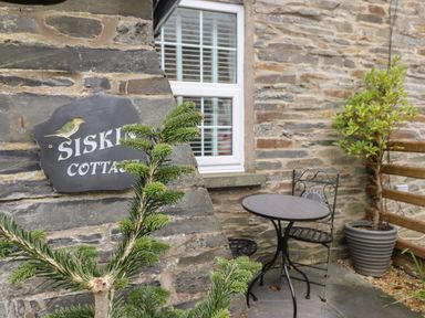A small outdoor seating area with a round metal table and two chairs next to a stone wall with a window and a sign reading Siskin Cottage at Siskin in Dinas Mawddwy near Dolgellau