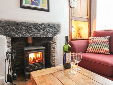 A fireplace with a wood stove burning and fire tools next to a wooden table with a bottle of wine and two glasses beside a red sofa with a patterned cushion at Siskin in Dinas Mawddwy near Dolgellau