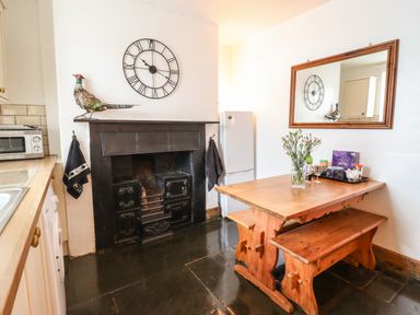 A kitchen with a wooden dining table and bench beside a black fireplace with a clock and decorative bird on the mantel at Siskin in Dinas Mawddwy near Dolgellau