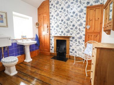 A bathroom with wooden floor, white sink, toilet with patterned seat, floral wallpaper, and wooden cabinets at Siskin in Dinas Mawddwy near Dolgellau
