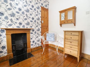 A room with a fireplace wallpapered wall a white metal chair with a blue cushion wooden chest of drawers and a wall cabinet at Siskin in Dinas Mawddwy near Dolgellau