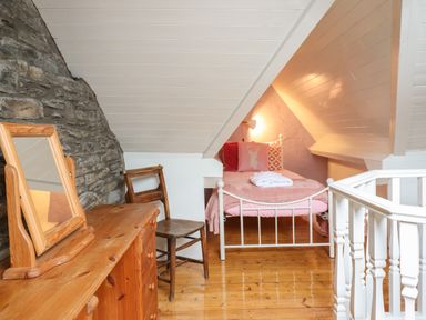A small bedroom with a single bed with pink bedding a wooden dresser with a mirror a wooden chair and exposed stone wall at Siskin in Dinas Mawddwy near Dolgellau