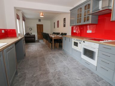 A long kitchen with grey cabinets and red backsplash a wooden dining table with black chairs and a brown leather armchair at Dumbuie in Haltwhistle