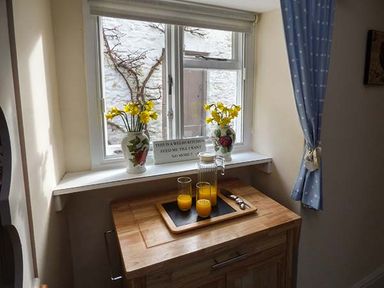 A window with two vases of yellow flowers and a wooden cabinet with a tray of orange juice glasses at Rhydlandgoed Stepaside near Saundersfoot