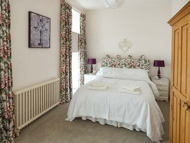 A bedroom with a bed with floral headboard and white bedding two purple lamps on white bedside tables floral curtains and a wooden wardrobe at Rhydlandgoed in Stepaside near Saundersfoot