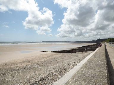 A beach with a pebble shore and wooden groynes next to a concrete sea wall and a road with cars at Rhydlandgoed Stepaside near Saundersfoot