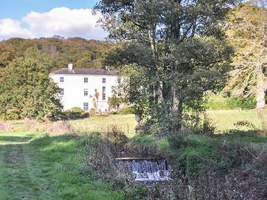 A large white house surrounded by trees and greenery with a small waterfall in the foreground at Rhydlandgoed Stepaside near Saundersfoot