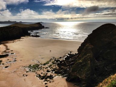 A sandy beach with rocky cliffs and two people walking near the shore at Sunset View in Porth