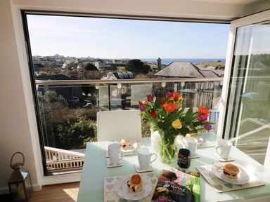 A dining table set with cups and scones next to a vase of flowers by an open window overlooking houses and the sea at Sunset View in Porth