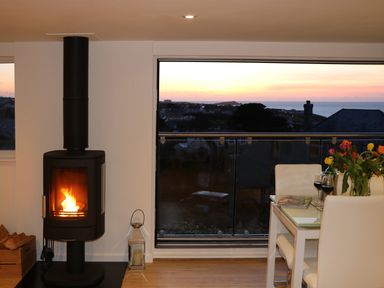 A room with a wood-burning stove next to a glass dining table with flowers and wine glasses overlooking a sunset at Sunset View in Porth
