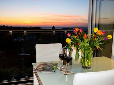 A glass table with two glasses of red wine a vase of flowers and a magazine on a balcony at Sunset View in Porth
